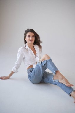 Beyaz gömlekli ve yırtık kot pantolonlu esmer kadın portresi. Yerde oturan genç bir kadın, beyaz stüdyo geçmişi. 