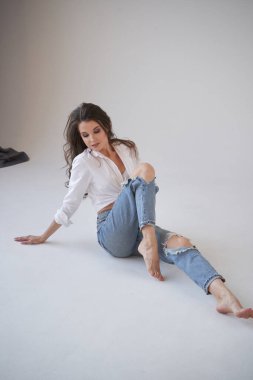Beyaz gömlekli ve yırtık kot pantolonlu esmer kadın portresi. Yerde oturan genç bir kadın, beyaz stüdyo geçmişi. 
