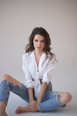 Beyaz gömlekli ve yırtık kot pantolonlu esmer kadın portresi. Yerde oturan genç bir kadın, beyaz stüdyo geçmişi. 