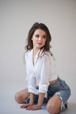 Beyaz gömlekli ve yırtık kot pantolonlu esmer kadın portresi. Yerde oturan genç bir kadın, beyaz stüdyo geçmişi. 