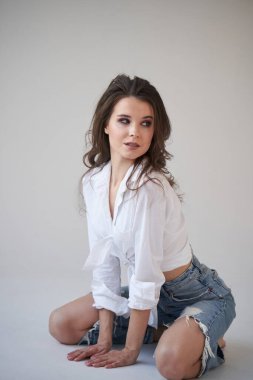 Beyaz gömlekli ve yırtık kot pantolonlu esmer kadın portresi. Yerde oturan genç bir kadın, beyaz stüdyo geçmişi. 