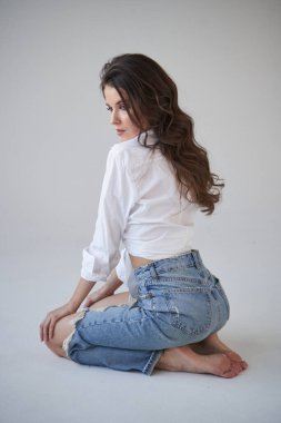 Beyaz gömlekli ve yırtık kot pantolonlu esmer kadın portresi. Yerde oturan genç bir kadın, beyaz stüdyo geçmişi. 
