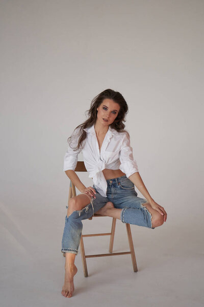 Портрет брюнетки в белой рубашке и разорванных синих джинсах. Молодая женщина сидит на деревянном стуле, белый студийный фон 