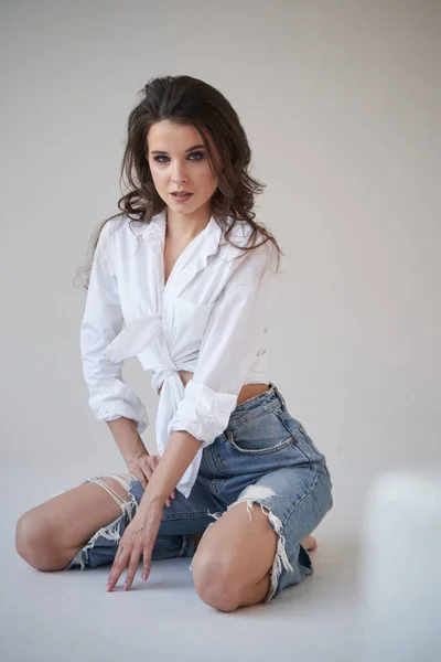 Portrait of beauty brunette in white shirt and ripped blue jeans. Young woman sitting on floor, white studio background