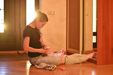 Kadın koridorda küçük kızına saç stili yapıyor. Güneş ön kapıdan içeri giriyor ve arka plan ışığı yaratıyor. Sıcak tonlu