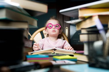 Çok sayıda kitap ve eğitim materyali arasında oyuncak gözlüklü bir çocuk bir masada oturuyor.