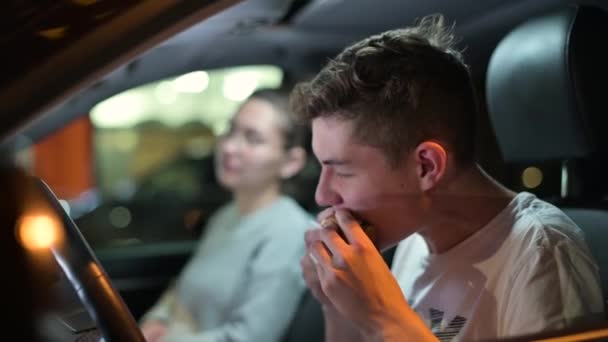 Fiatal srác, tinédzser, este hamburgert eszik a kocsiban a parkolóban. — Stock videók