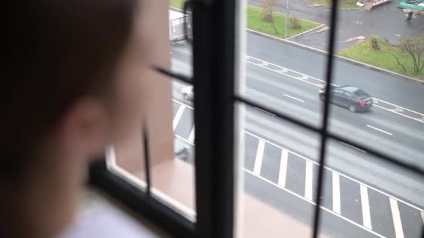 Uma jovem mulher olha pela janela para carros que passam na estrada perto de sua casa — Vídeo de Stock