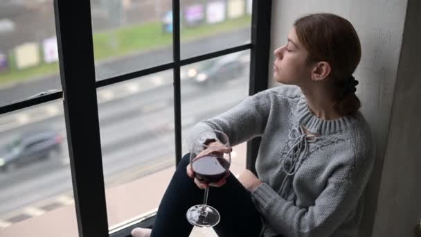 En ung kvinna med ett glas vin sitter på fönsterbrädan i en stadslägenhet — Stockvideo