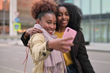 İki gülümseyen genç zenci kadın turist şehir manzaralarının arka planında selfie çekiyor.