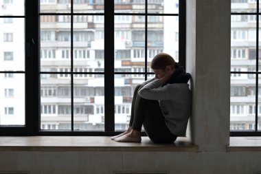 Hasta görünümlü genç bir kadın, bir şehir dairesinin pencere eşiğinde üzgün. Bir şehir sakininin sonbahar bunalımı