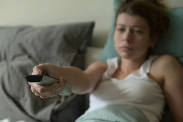 电视控制面板是在一个疲倦的 生病的年轻女子手中 她正在更换频道 年轻女人生病了 无聊了 就在床上呆着 — 图库照片