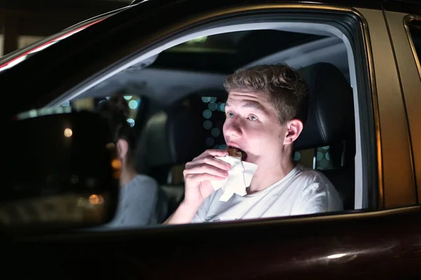 Young man eating burger in the car driving late at night in the parking lot. Close up, view through window