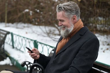 Saf gri saçlı bir adam parkta bir bankta oturur ve video bağlantısıyla konuşur. Telefonla konuşan sakallı olumlu bir adam.