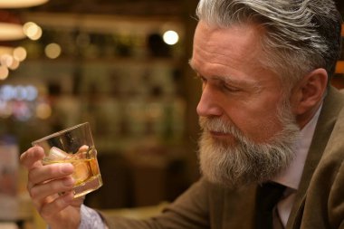 Adam elindeki viski bardağına bakıyor. Yetişkin bir adam restoranda içiyor.