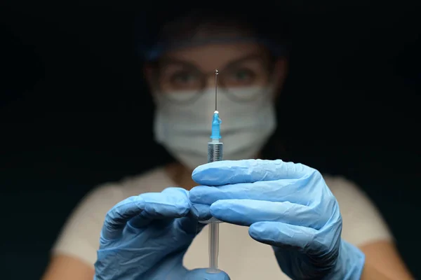 a nurse or doctor in a mask and gloves raises a syringe with a medicine or vaccine in the dark. The symbol of getting rid of the disease in the hands of a doctor