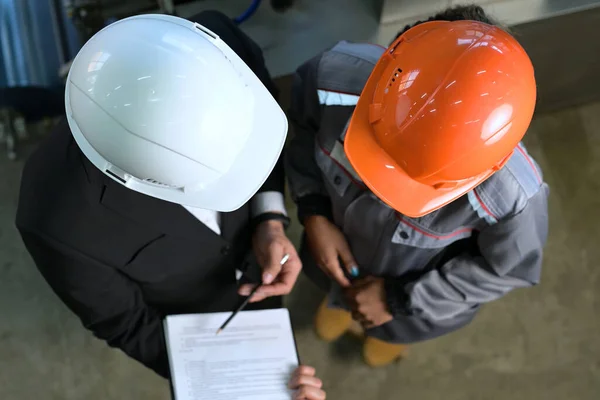 Werknemer en baas in oranje en witte helmen praten over werk in productie Rechtenvrije Stockafbeeldingen