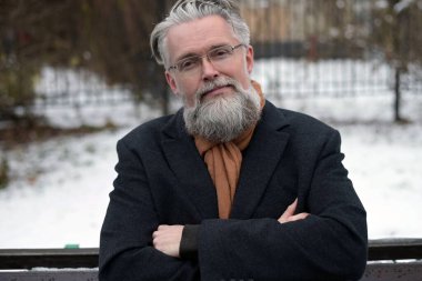 Yetişkin, gri saçlı, gözlüklü bir adam bankta oturur ve kameraya bakar.