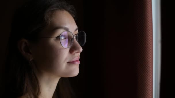 Mujer joven en gafas se ve pensativamente por la ventana desde una habitación oscura — Vídeo de stock