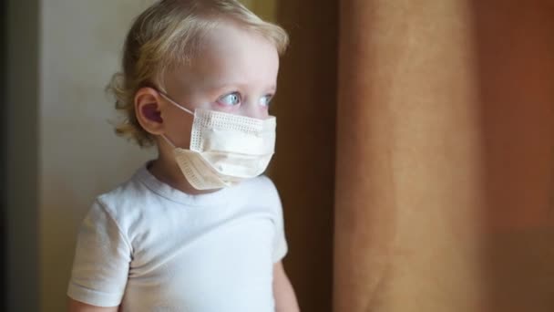 Petite fille dans un masque médical de protection se tient près des rideaux de fenêtre. — Video