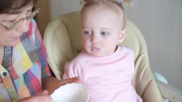 En liten flicka som äter yoghurt från en sked. Mormor matar sitt barnbarn — Stockvideo