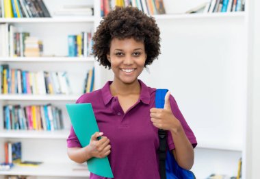 Sırt çantası ve evrakları olan Afro-Amerikalı kız öğrenci üniversitenin sınıfında baş parmağını gösteriyor.