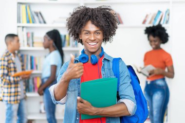 Afro-Amerikan erkek üniversite öğrencisi üniversitenin kütüphanesinde baş parmağını gösteriyor.