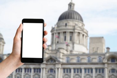 Hand holding mobile phone with blank screen and historic building at Liverpool at Great Britain