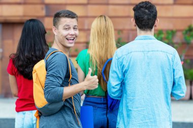 Alman erkek öğrenci, üniversitenin kampüsünde diğer öğrencilerle baş parmağıyla boy gösteriyor.