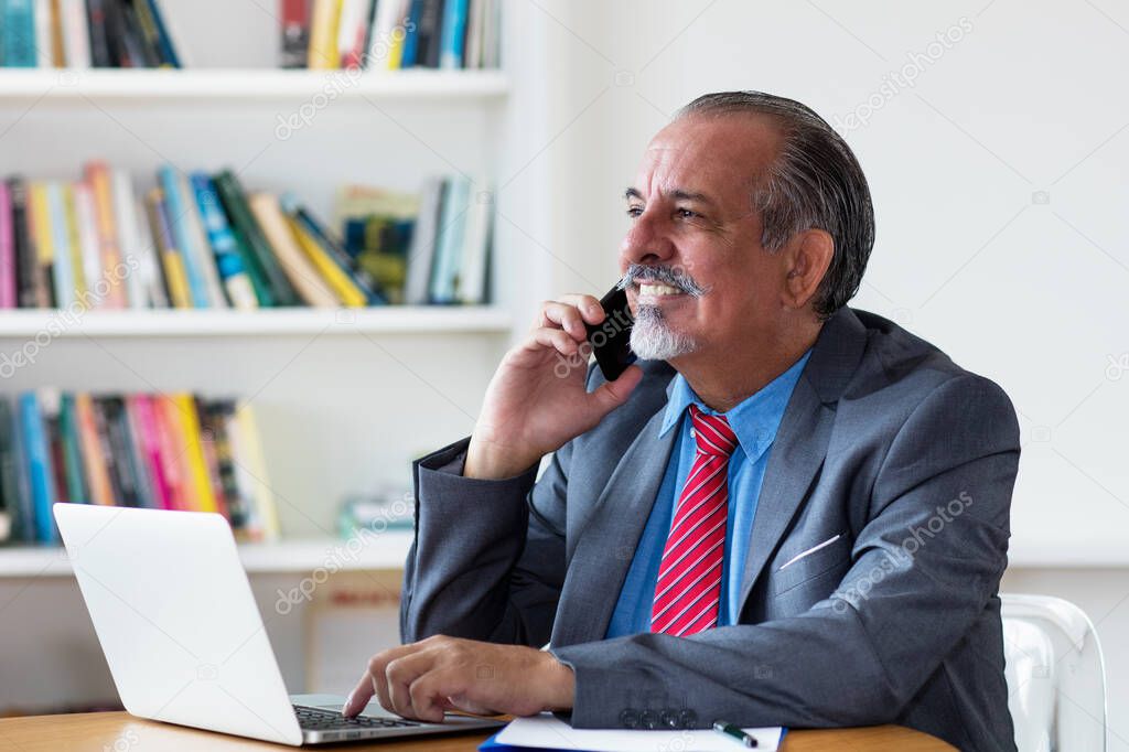 Mexican senior businessman talking with customer at phone at desk at office