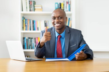 Zeki Afro-Amerikan olgun iş adamı şirket bürosundaki masada baş parmağını gösteriyor.