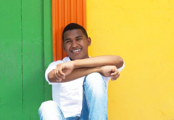 Laughing caribbean guy sitting in front of a colorful wall — Stock Photo, Image