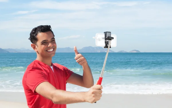 Feliz chico latino en la playa hablando de una foto con teléfono y selfie —  Fotos de Stock