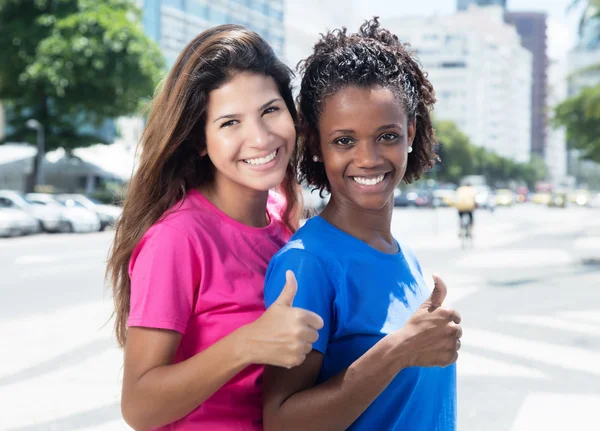 Two african and caucasian women showing thumb in the city — Stock Photo, Image