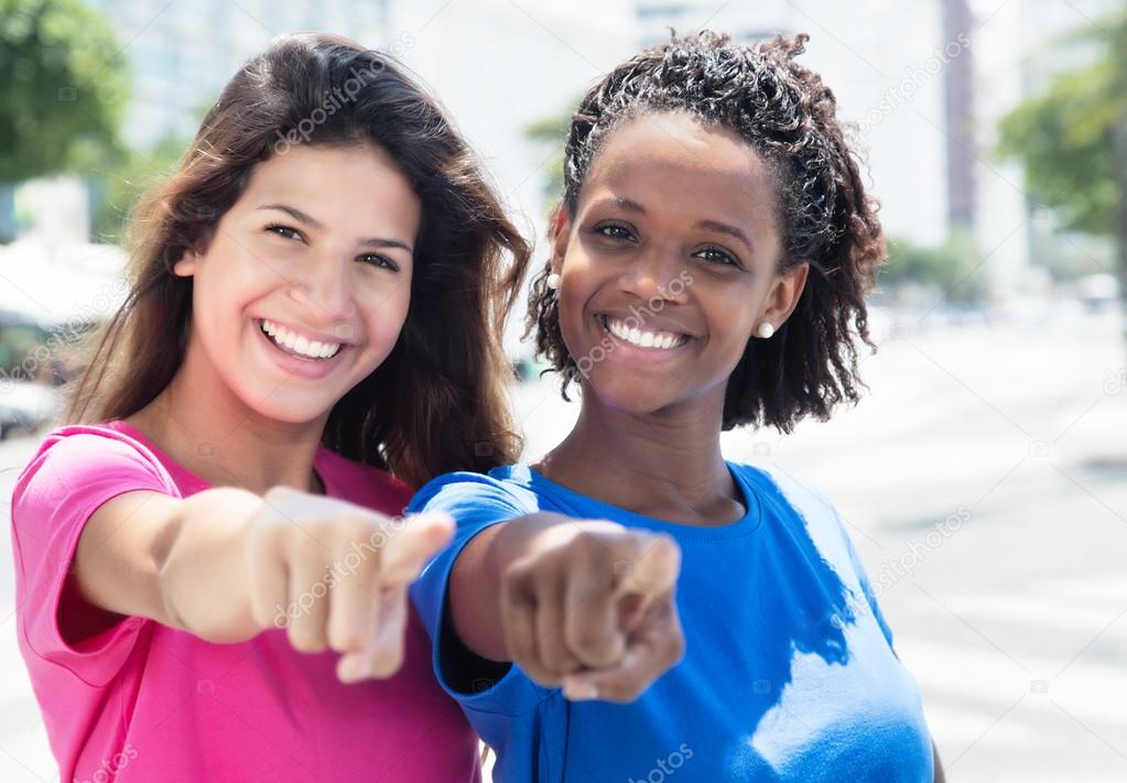 Two african and caucasian women pointing at camera in the city