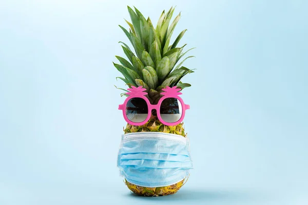 COVID Concepto de viaje. Piña en gafas de sol y máscara médica sobre fondo azul. Piña en gafas de sol rosas y mascarilla - Nuevo viaje normal — Foto de Stock