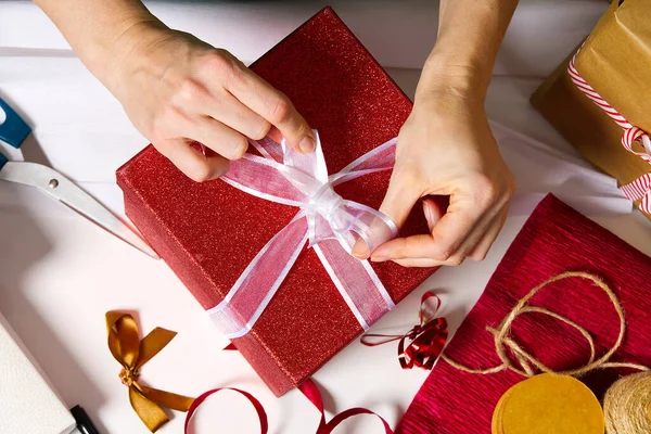 Kadın elleri kırmızı bir kutuda beyaz kurdeleli bir yılbaşı hediyesi paketliyor. Hediye paketi. Yılbaşı paketleme ve paketleme hizmeti konsepti