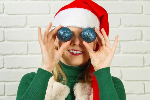 Christmas fun. woman in santa red hat holding Christmas balls in front of her eyes and smiling assign white brick wall — Stock Photo, Image