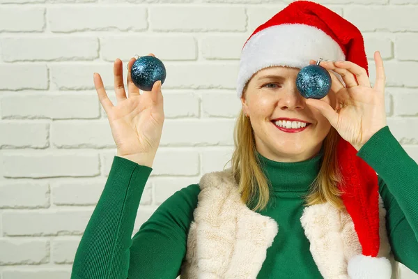 Karácsonyi móka. nő télapó piros kalap tartja karácsonyi golyók előtt a szeme és mosolygós hozzárendel fehér tégla fal — Stock Fotó