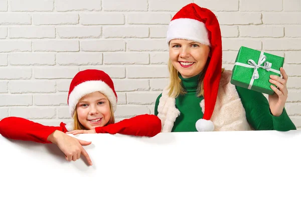 Anya és lánya leskelődnek az üres táblák mögül, és fehér téglafalat jelölnek ki. Törpe lány, másold a helyet. Karácsonyi koncepció — Stock Fotó