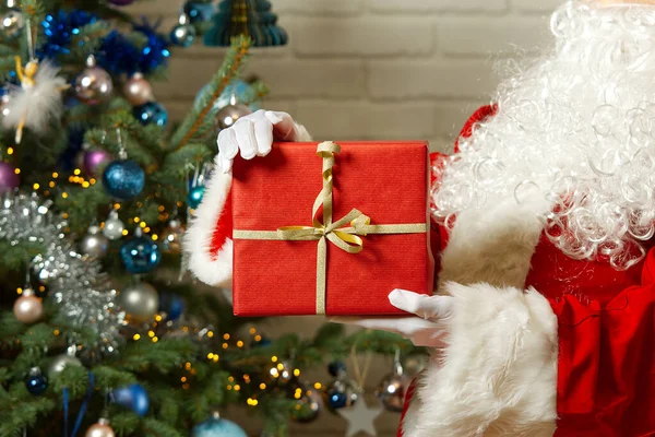 Santa Claus manos sostiene caja de regalo roja frente al árbol de Navidad. — Foto de Stock