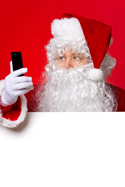 Papai Noel segurando o telefone celular na mão enquanto estava atrás da placa branca em branco. espaço de cópia. Texting Papai Noel. — Fotografia de Stock