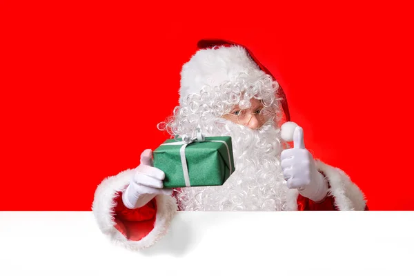 Santa Claus sostiene la caja de regalo verde en la mano mientras está de pie detrás de la pizarra blanca en blanco. espacio de copia. — Foto de Stock