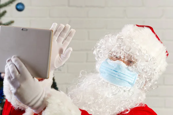 Hombre en traje de Santa Claus y máscara de gripe con una Tablet Computer. felicitaciones de santa claus a través de la tableta. — Foto de Stock