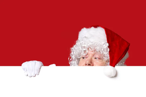 Der Weihnachtsmann schaut hinter der leeren weißen Tafel auf rotem Hintergrund mit Kopierraum hervor — Stockfoto