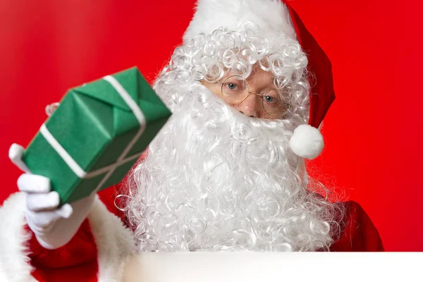 Papai Noel segurando caixa de presente verde na mão enquanto estava atrás da placa branca em branco. espaço de cópia. — Fotografia de Stock