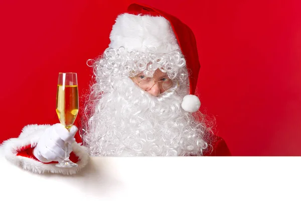 Papai Noel segurando copo de vinho espumante na mão enquanto estava atrás da tábua branca em branco. espaço de cópia. — Fotografia de Stock