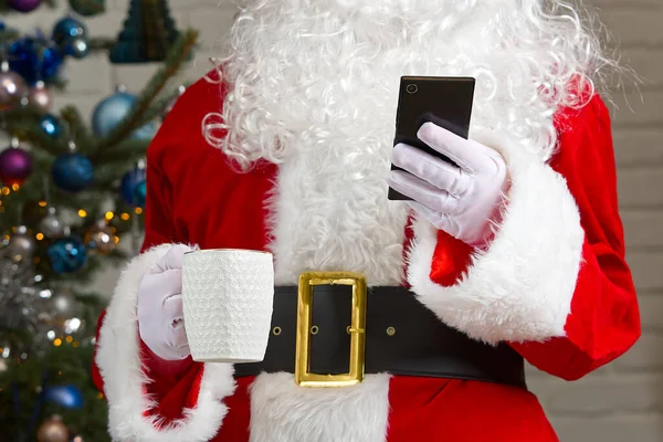 Santa claus holding white mug with coffee and smartphone in his hand, close-up. mobile Phone And Coffee mug in santa hands. — Stock Photo, Image