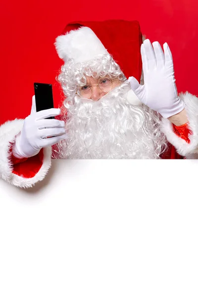 Santa Claus sosteniendo el teléfono móvil en la mano mientras está de pie detrás de la pizarra blanca en blanco. espacio de copia. Santa Claus mensajes de texto. — Foto de Stock