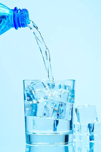 Pouring fresh clean drinking water from blue plastic bottle into glass on blue background close up